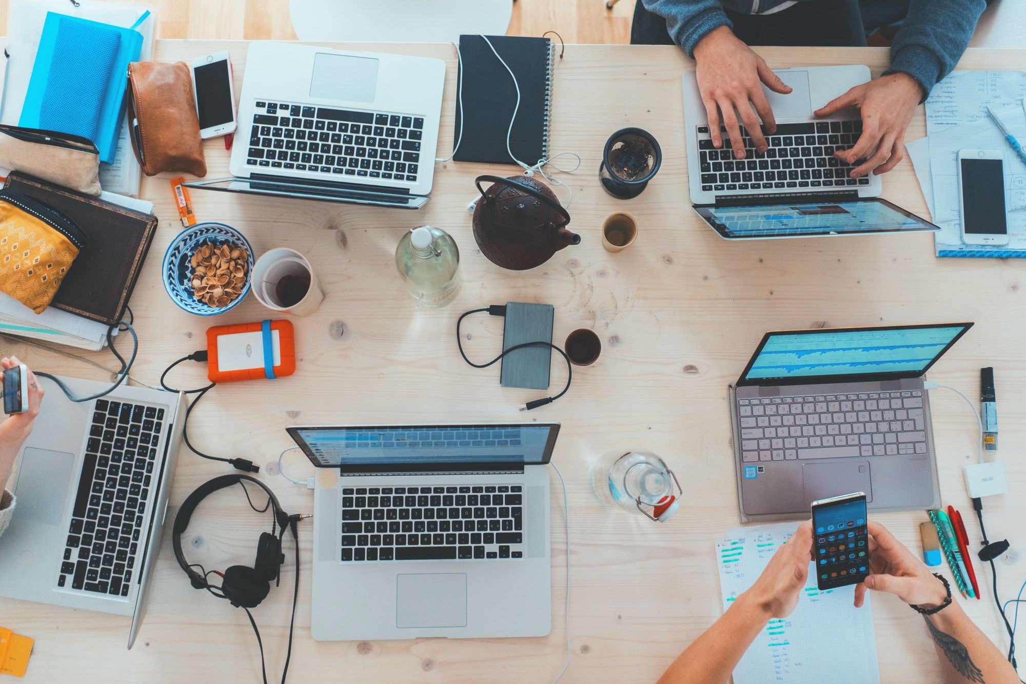 A flat-lay photo of a wood desk with five laptops, and three people using phones or typing.